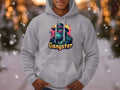 A person in a gray hoodie stands outdoors against a snowy backdrop. The medium-heavy fabric hoodie by Garment Graphics features a colorful gangster graphic of a blue gorilla in a suit, smoking, with Gangster printed below, highlighting its smokey cool style.