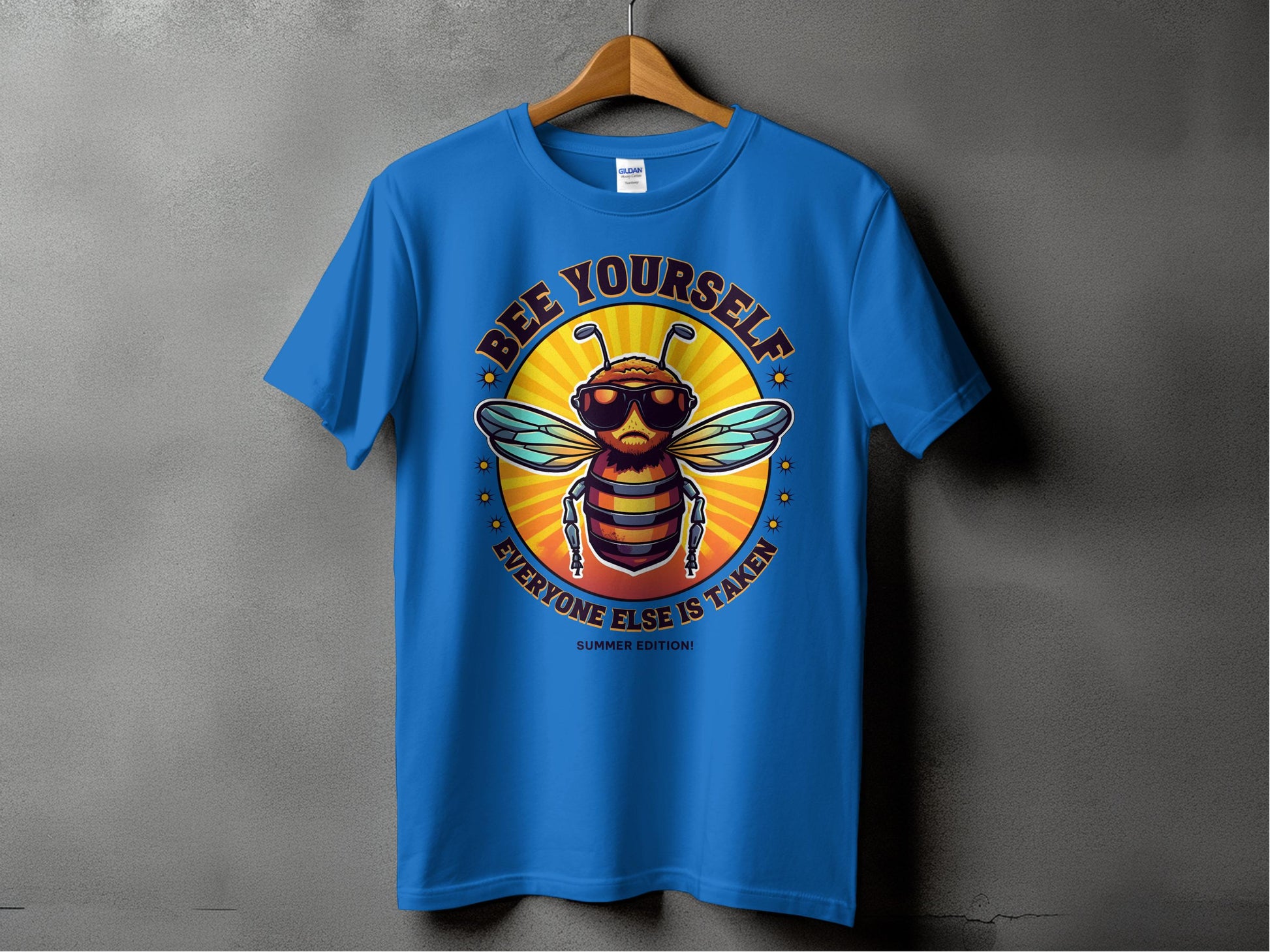A blue T-shirt from Garment Graphics Summer Edition hangs on a gray wall, featuring a cool bee with sunglasses and a jacket. The text reads Bee Yourself above and Everyone Else Is Taken below. Available in family sizing, its perfect for spreading the buzz!.