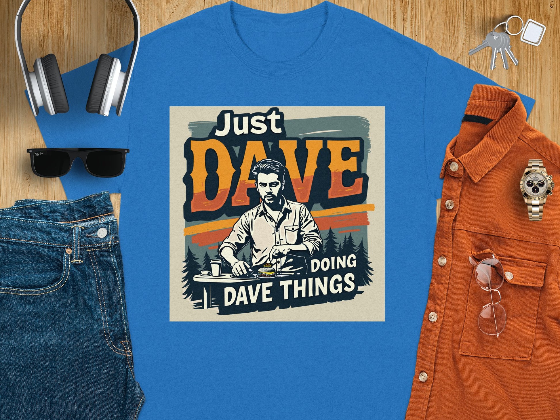 A blue classic fit graphic T-shirt from Garment Graphics features Just Dave Doing Dave Things with a man at a table, styled on wood with a watch, orange shirt, keys, headphones, sunglasses, and denim pants for an effortlessly cool vibe.