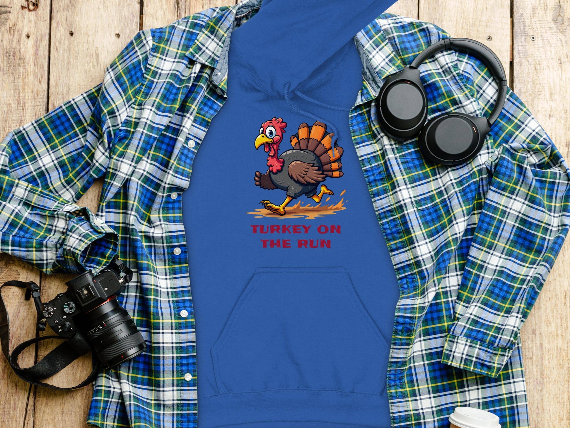 A blue Garment Graphics Turkey on the Run hoodie with a cartoon turkey graphic sits on a wooden surface. Nearby are a blue and green plaid shirt, camera, headphones, and lidded cup—ideal for fans of funny hoodies and photography.