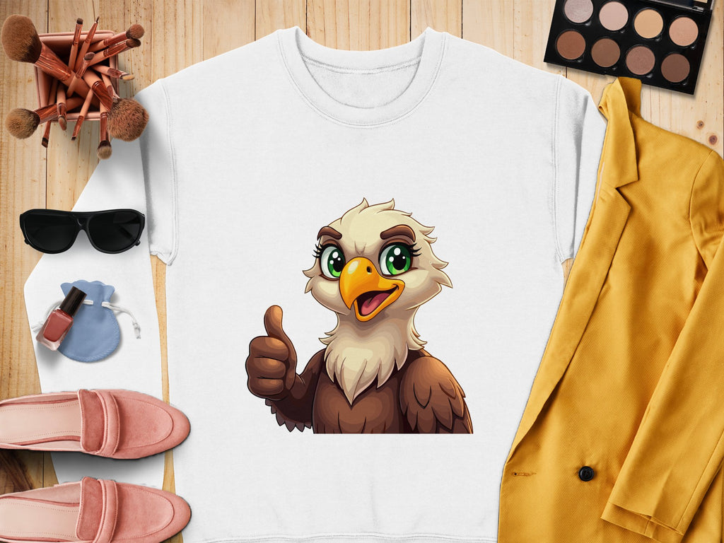 A flat lay highlights a white Garment Graphics sweatshirt with a cheerful cartoon eagle giving a thumbs-up. Its surrounded by pink shoes, sunglasses, makeup brushes and palette, a yellow coat, and a small blue pouch.