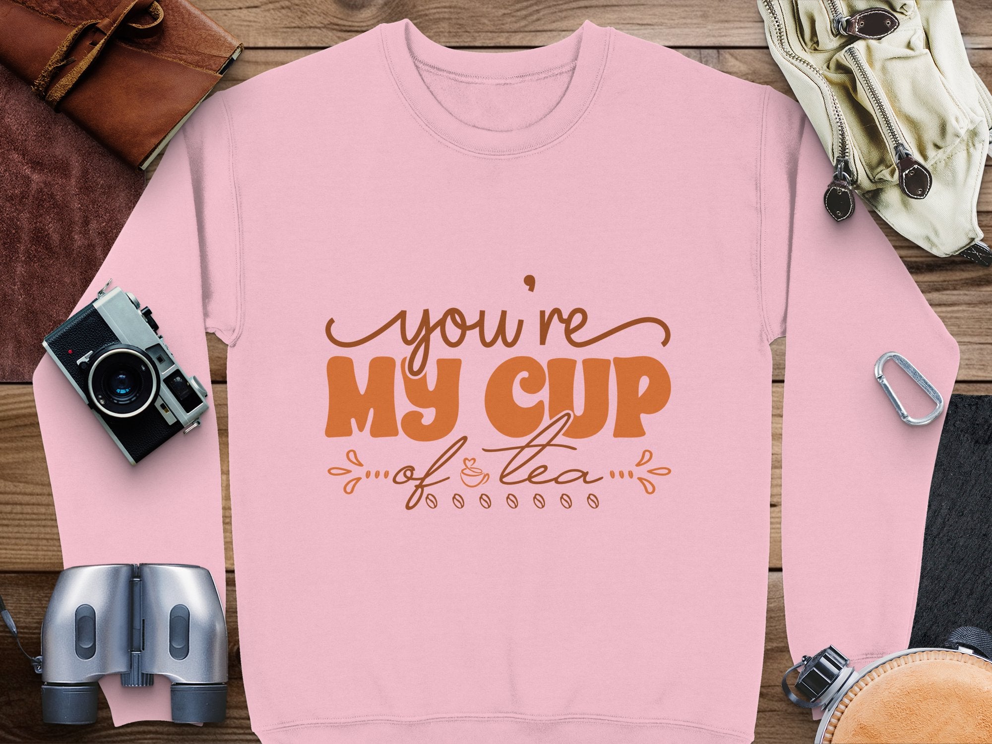 A cozy pink sweatshirt by Garment Graphics, in unisex sizing, features Youre My Cup of Tea in brown lettering with decorative details. Surrounded by a camera, binoculars, leather bag, goggles, and carabiners when laid flat—ideal for adventure lovers.