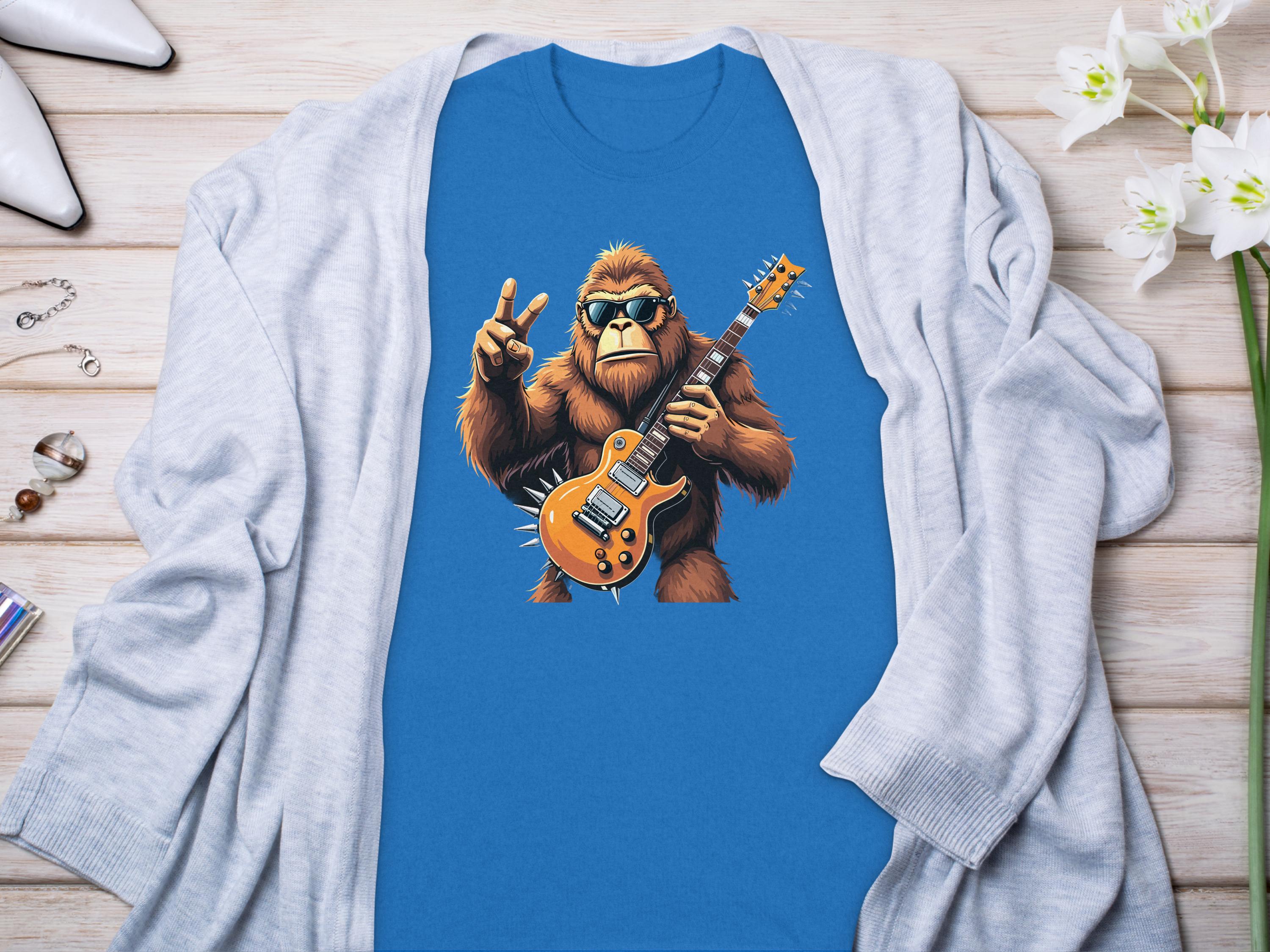 A blue Garment Graphics T-shirt for music lovers features a gorilla with an electric guitar and peace sign, paired with a light gray cardigan. White shoes, jewelry, and white flowers adorn the wooden surface nearby.