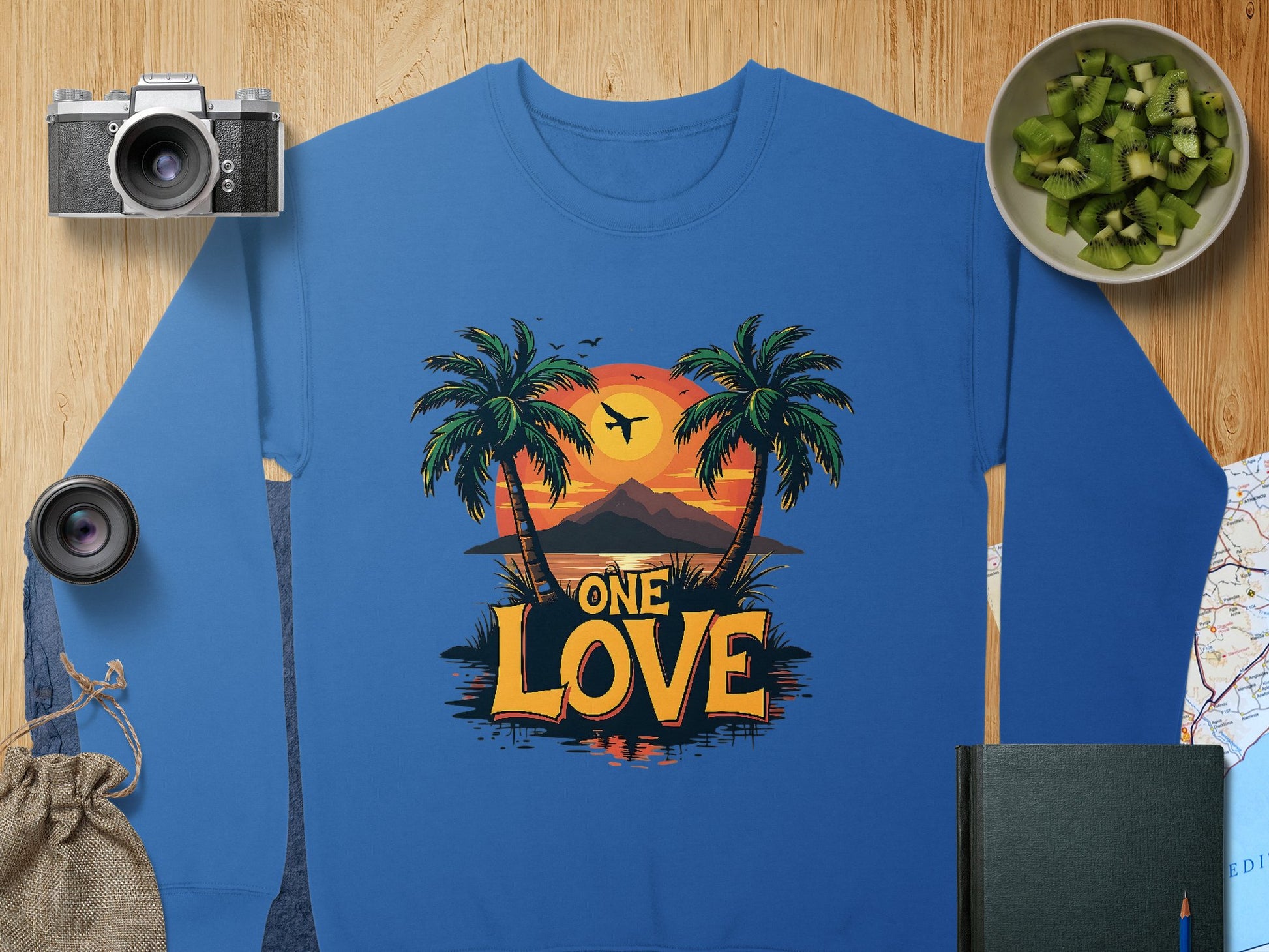 Garment Graphics blue sweatshirt features a vibrant One Love Sunset print with palm trees and an airplane. Styled on a wooden surface, its paired with a camera, lens, green bowl, drawstring bag, closed book, and map—ideal for tropical dreamers.