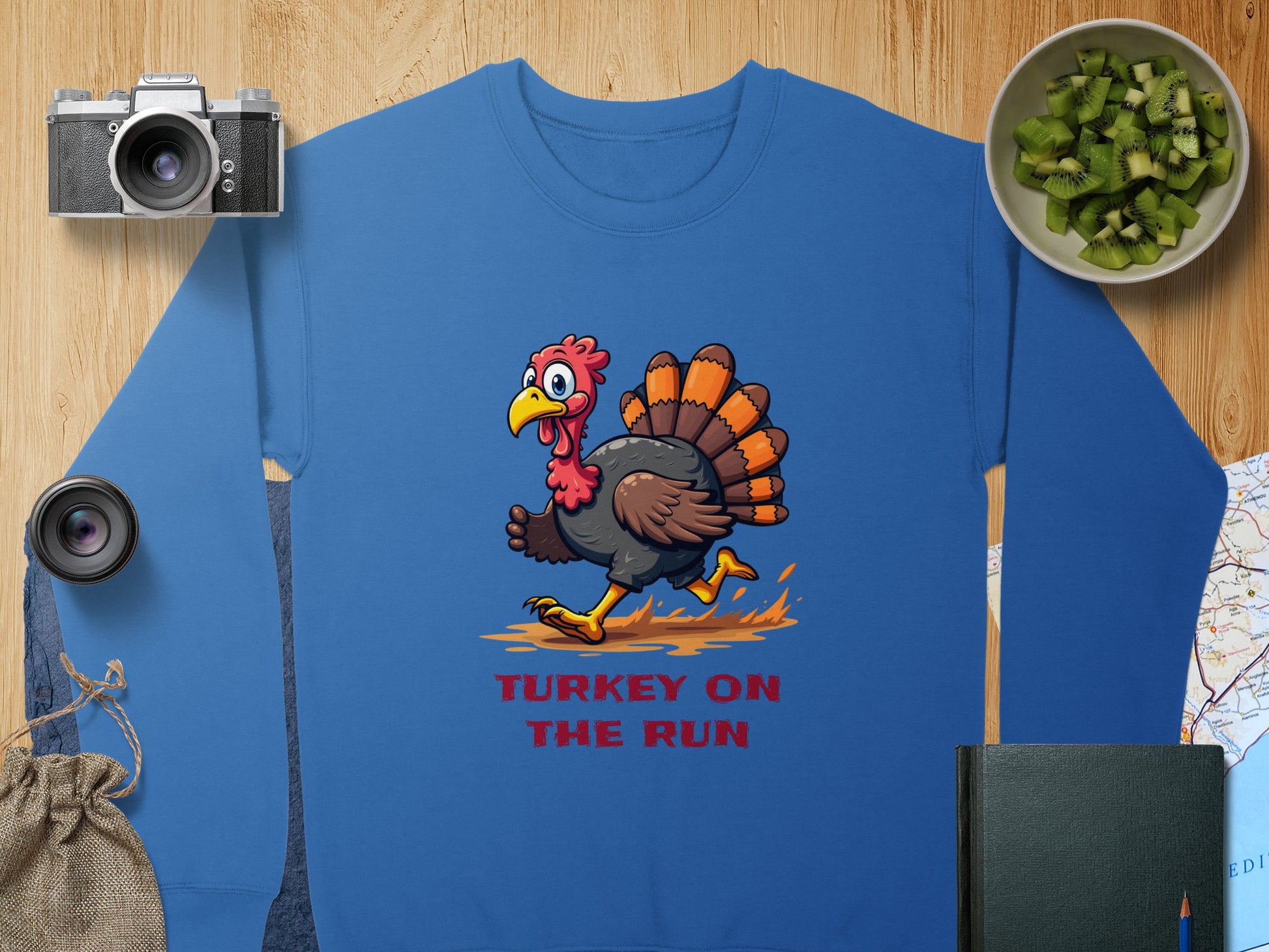 Garment Graphics blue unisex sweatshirt showcases a comedic cartoon turkey design with Turkey on the Run underneath. Its laid out on a wooden surface with items like a camera, lens, bag, sliced cucumber bowl, book, and map surrounding it.