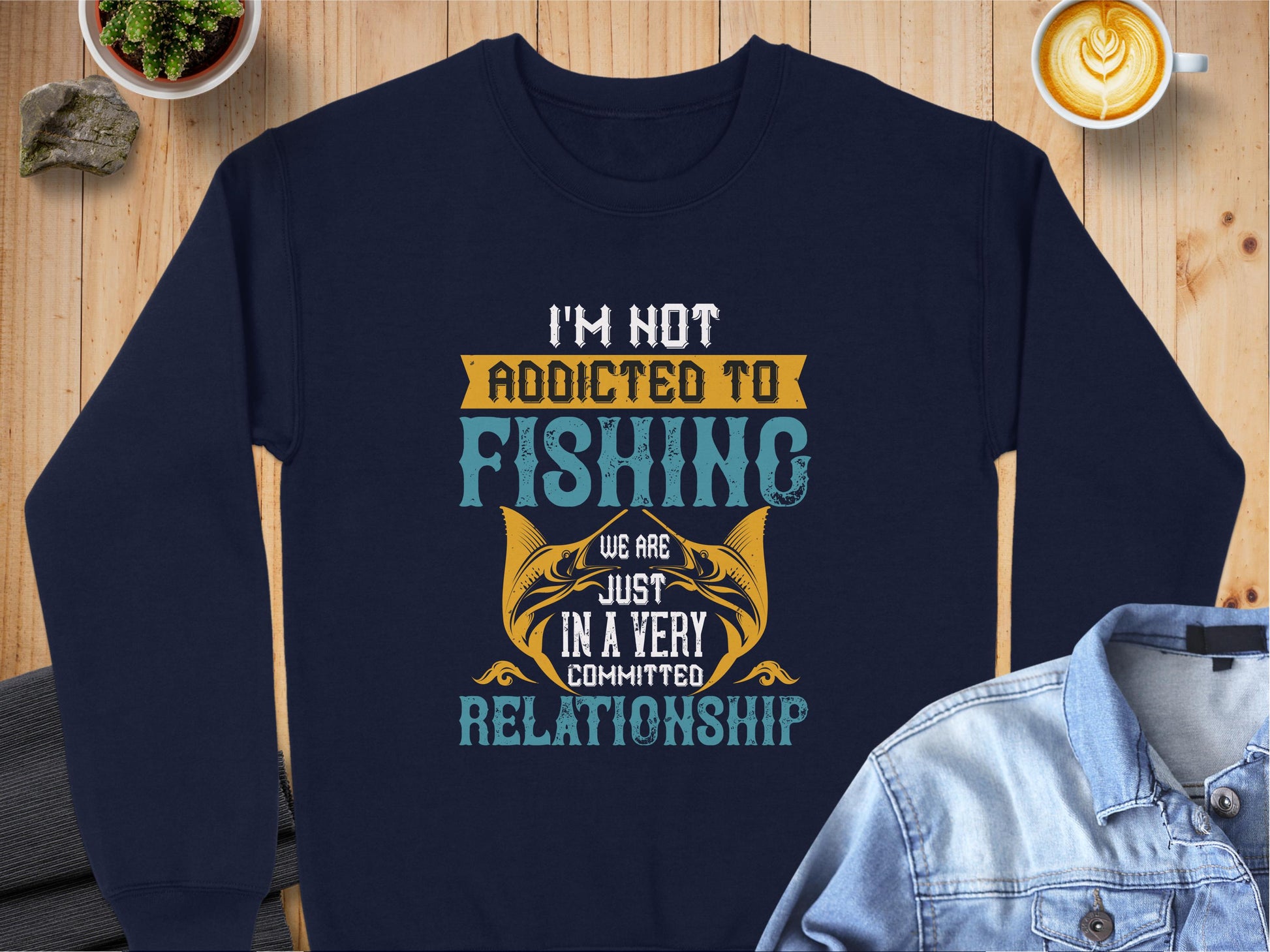 A Garment Graphics navy blue unisex sweatshirt lies on the table, featuring a fun fishing quote: Im not addicted to fishing, we are just in a very committed relationship. Alongside it are a latte, denim shirt, plant, and rock.