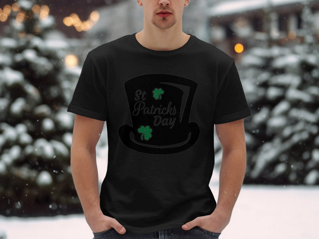 A man stands in a snowy landscape wearing a black Garment Graphics T-shirt, featuring a top hat design with green shamrocks and four-leaf clovers, celebrating St. Patricks Day.