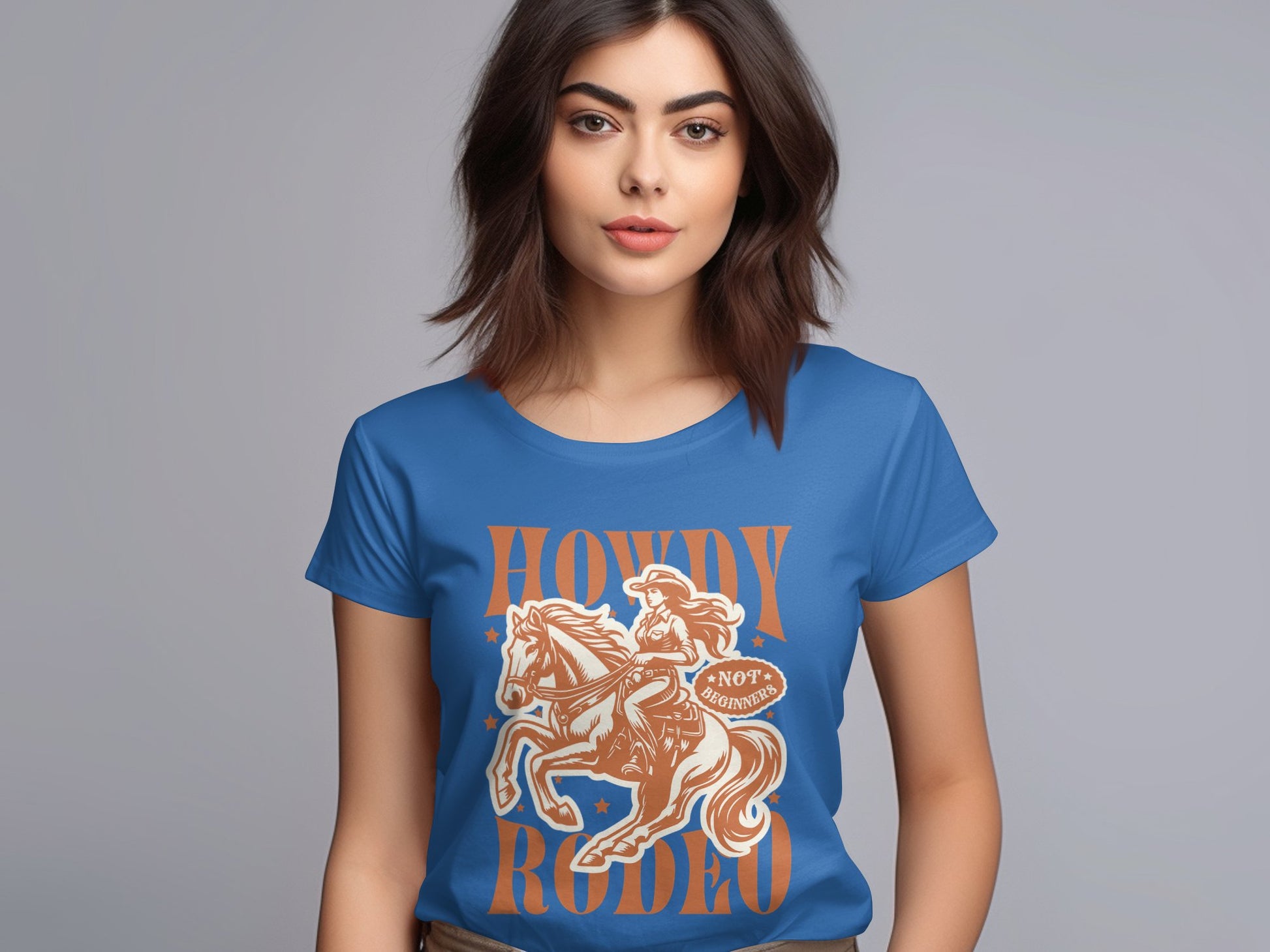 A woman with dark hair displays a blue Garment Graphics Rodeo T-Shirt in womens sizes, featuring a cowboy on horseback and the phrase Howdy Rodeo in vibrant orange, standing against a plain gray background.