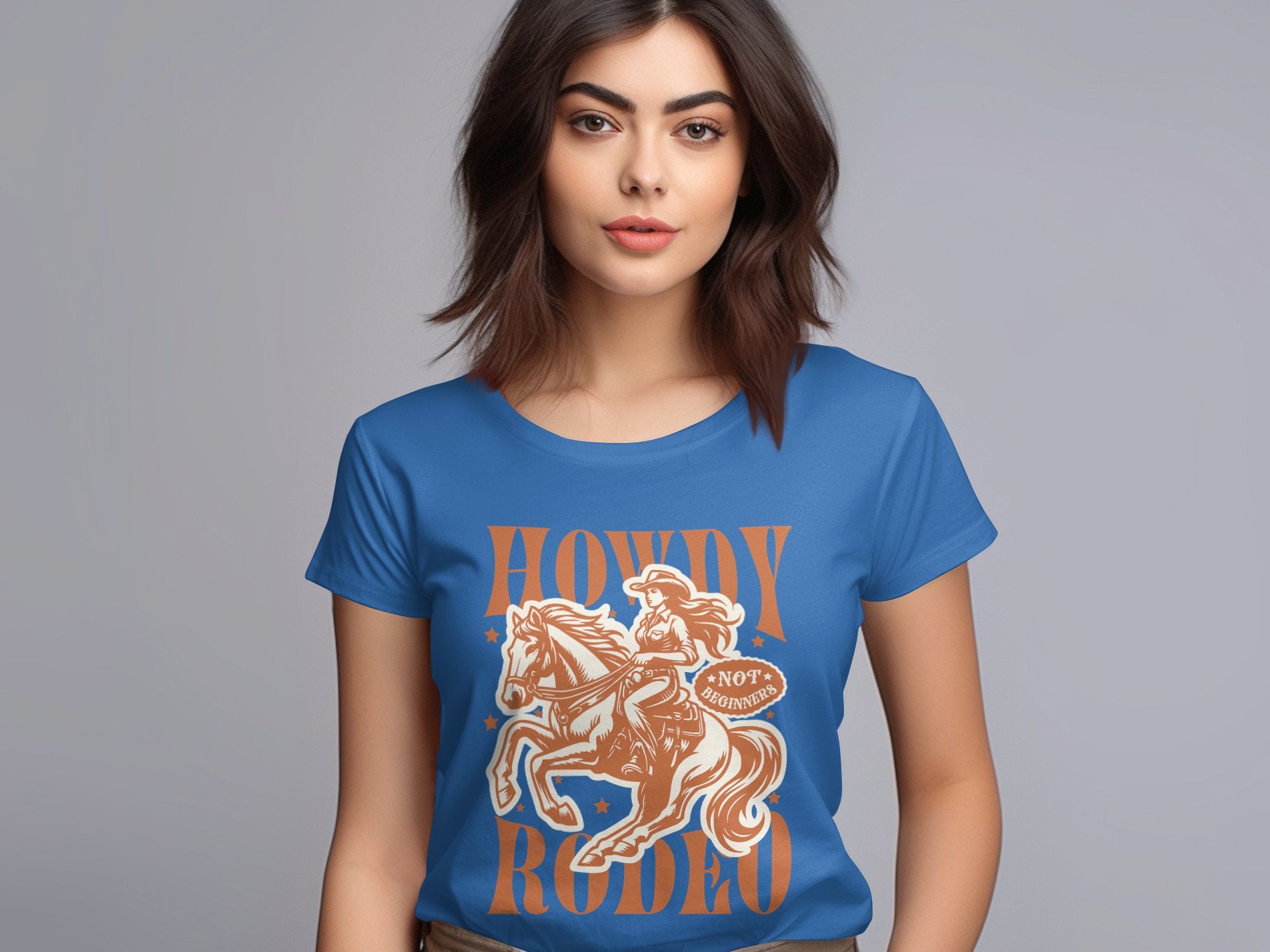 A woman with dark hair displays a blue Garment Graphics Rodeo T-Shirt in womens sizes, featuring a cowboy on horseback and the phrase Howdy Rodeo in vibrant orange, standing against a plain gray background.