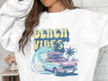 A person is wearing a white Garment Graphics sweatshirt featuring a classic car with surfboards, cruising by waves and palm trees, with BEACH VIBES in bold retro-style letters.