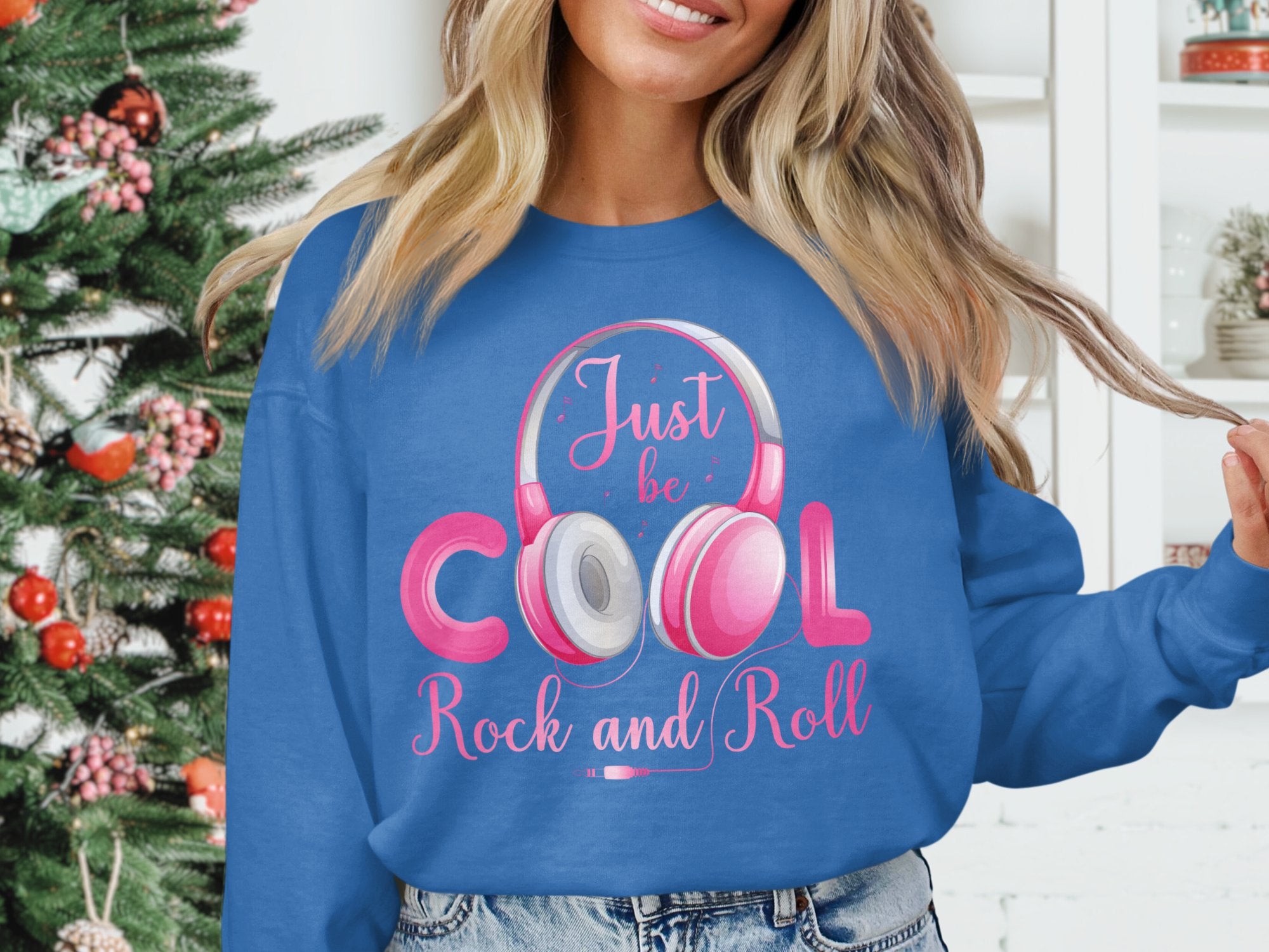 A person stands before a decorated Christmas tree wearing a Garment Graphics blue sweatshirt with pink and white accents, paired with Rock and Roll headphones. Just be Cool captures the music lovers spirit, while white shelves filled with decorations complete the background scene.