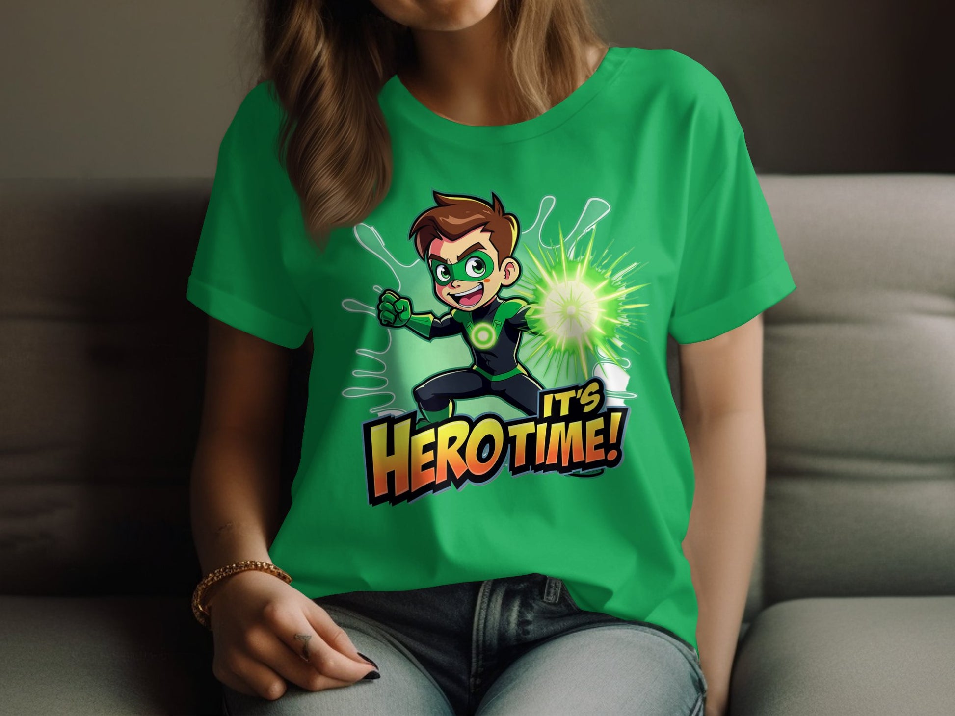 A person sits on a sofa wearing Garment Graphics green t-shirt featuring a cartoon superhero with brown hair, a green suit, and a glowing hand. The shirt adds color with the words Its Hero Time! printed below.
