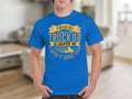 Someone wearing a blue T-shirt from Garment Graphics featuring the text: If hes got golf clubs in his truck or a camper in his driveway, dont hire him. The shirts camping humor stands out against what looks like a cozy, blurred living room background.