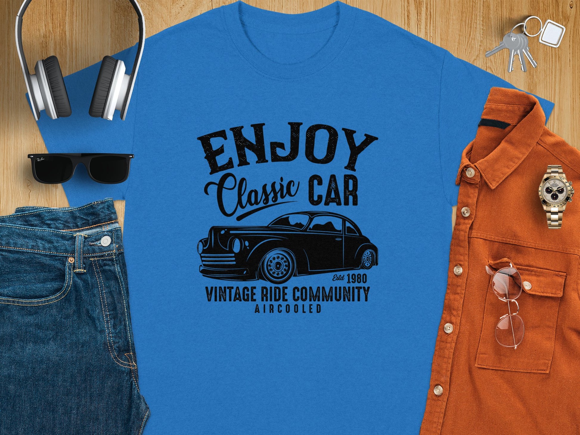 Flat lay featuring a blue Enjoy Classic Car T-shirt by Garment Graphics with a car graphic, ideal for car lovers, surrounded by headphones, sunglasses, jeans, an orange shirt, watch, and keys on a wooden surface.