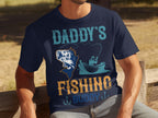 A fishing enthusiast unwinds on a wooden bench, wearing a navy graphic tee by Garment Graphics. The shirt displays Daddys Fishing Buddy with a colorful fish and someone casting from a boat illustration.