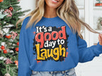 A person stands by a decorated Christmas tree wearing a Garment Graphics sweatshirt with Its a good day to Laugh in colorful text. The medium-heavy fabric offers comfort for all with unisex sizing. The background shows a white interior with shelves and decorative items.