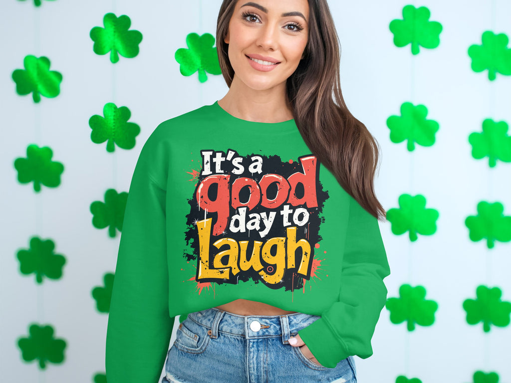 A person with long dark hair wears a green graphic sweatshirt from Garment Graphics reading Its a good day to Laugh in colorful letters. They stand before a shamrock-decorated wall, emphasizing the medium-heavy fabric ideal for unisex sizing.