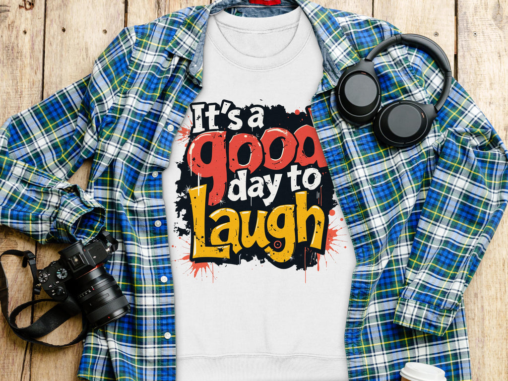 A flat lay captures the Garment Graphics white T-shirt stating, Its a Good Day to Laugh, layered over a blue and green plaid shirt. Its surrounded by essentials: a camera, headphones, and coffee cup on the wooden surface, highlighting the unisex styles relaxed charm.