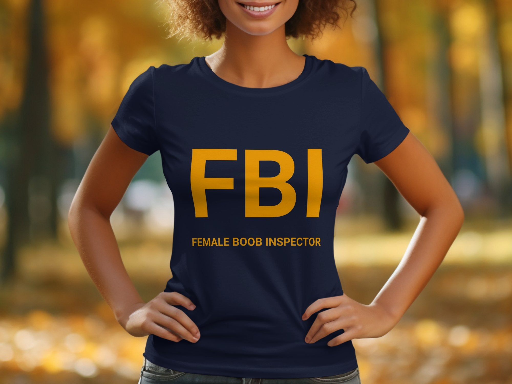 In a blurred autumn scene, someone wears a Garment Graphics navy blue FBI Female Boob Inspector T-shirt, humorously styled as a gag gift. Large yellow letters stand out above Female Boob Inspector, and womens sizing adds a fun twist.