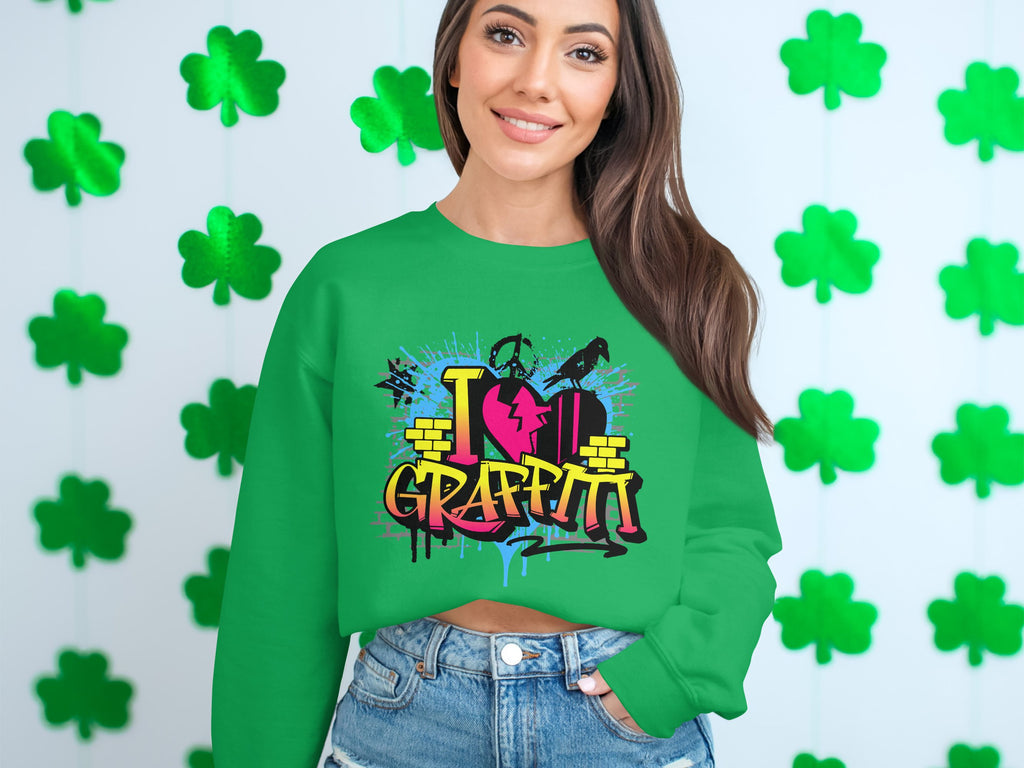 A woman stands against a shamrock-themed backdrop, wearing a Garment Graphics sweatshirt featuring a lively I Love Graffiti design. Her urban look is highlighted by long brown hair and denim shorts.