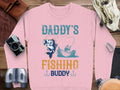 A Garment Graphics pink sweatshirt, ideal for fishing fans, is on a wooden surface. It shows a person fishing from a boat with a jumping fish and Daddys Fishing Buddy. Nearby are a camera, binoculars, and compass.