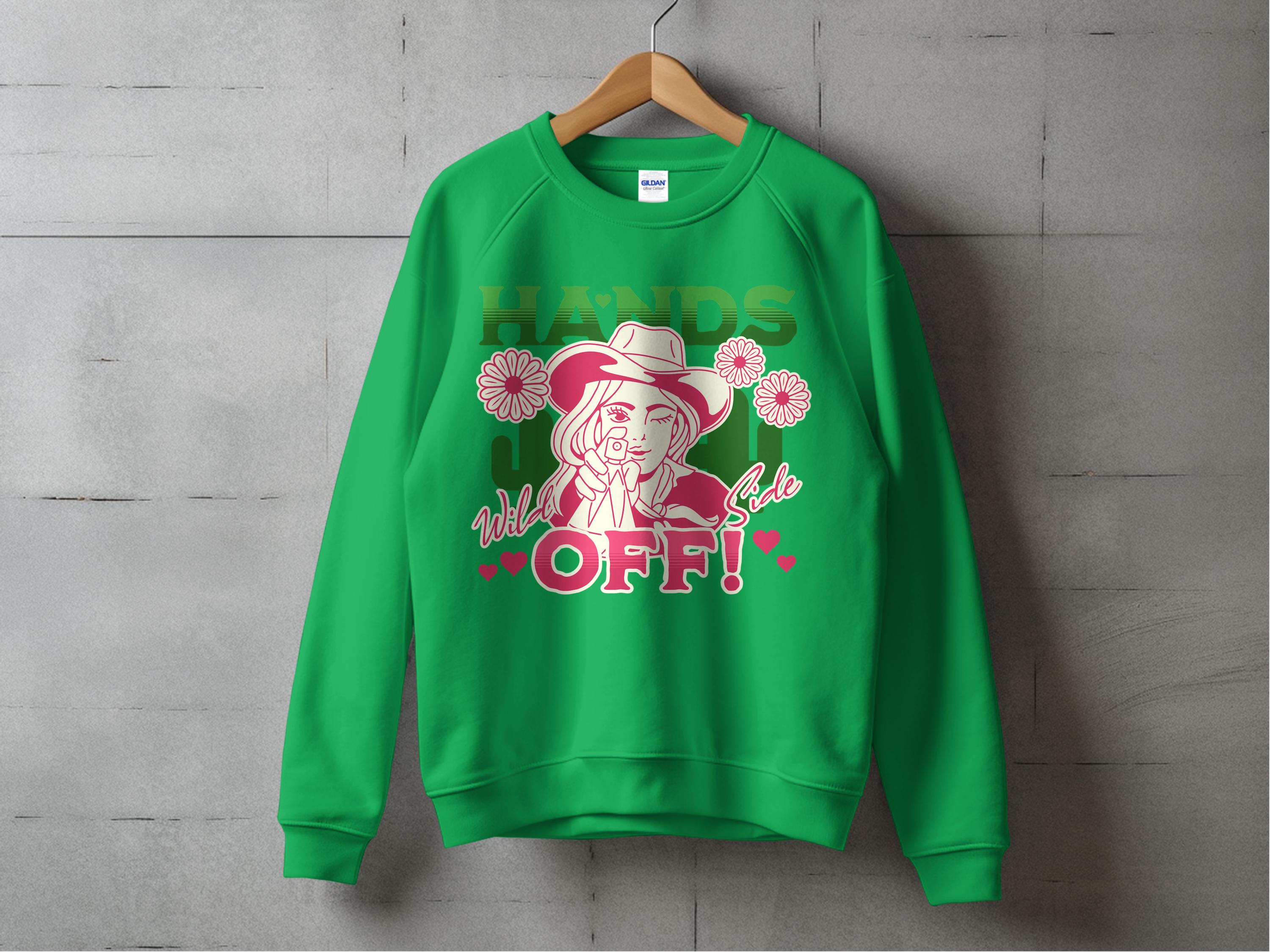 The Garment Graphics Wild Side Sweatshirt, in green and unisex sizing, hangs on a wooden hanger against a gray wall. It features bold graphics of a hatted figure with flowers and the phrase HANDS OFF! enhanced by pink and white flowers and hearts for an enchanting touch.