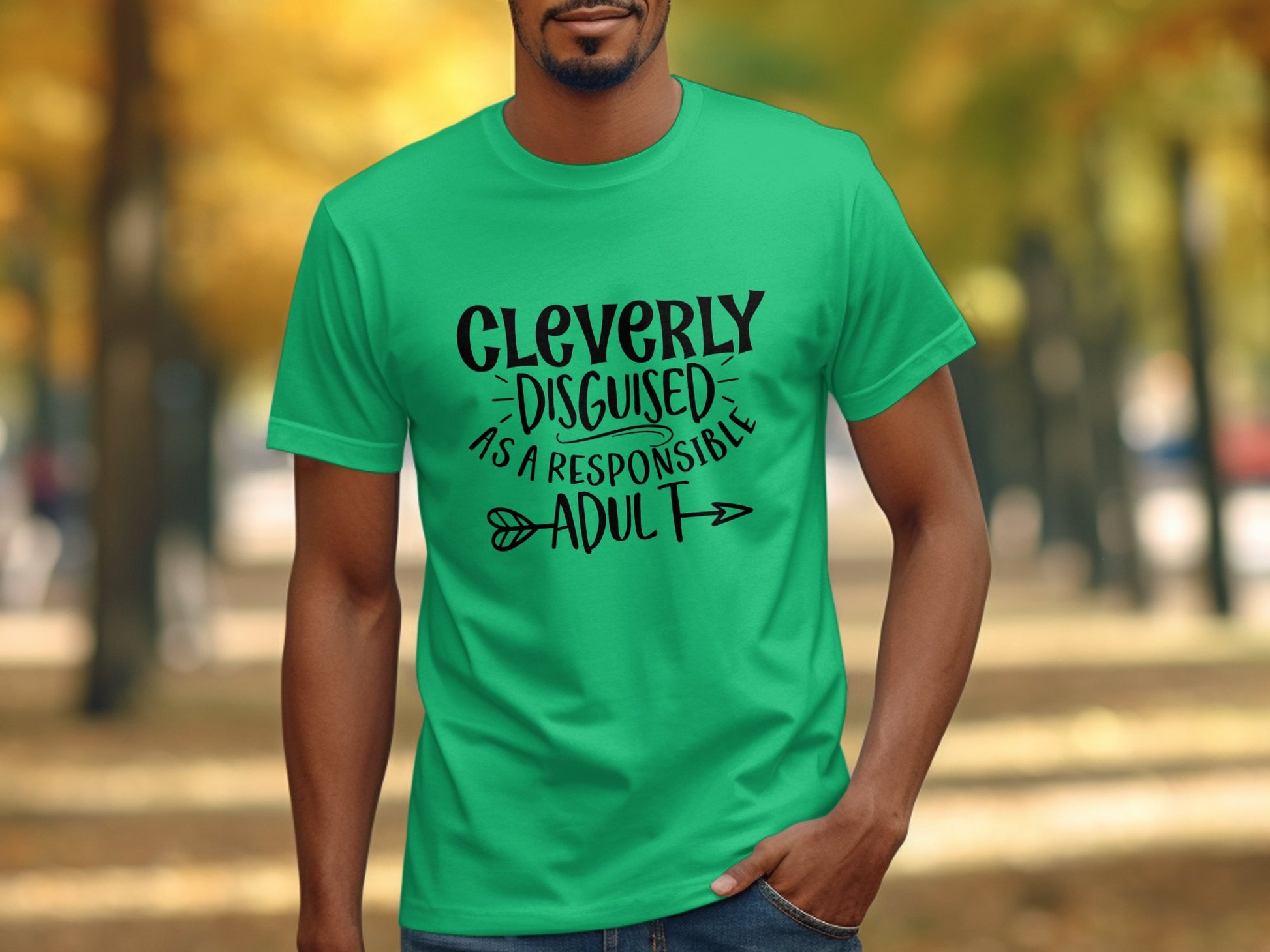 Wearing a classic fit green Garment Graphics shirt that reads Cleverly Disguised as a Responsible Adult, a person enjoys a casual outing against the backdrop of blurred autumn trees.