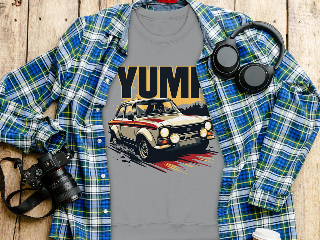 A gray t-shirt with a vintage rally car graphic and YUMI from Garment Graphics is on a wooden surface, paired with a blue and green plaid shirt echoing the YUMP style. Nearby are essentials like a camera, headphones, and lidded cup for the perfect casual outing.