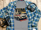 A gray t-shirt with a vintage rally car graphic and YUMI from Garment Graphics is on a wooden surface, paired with a blue and green plaid shirt echoing the YUMP style. Nearby are essentials like a camera, headphones, and lidded cup for the perfect casual outing.