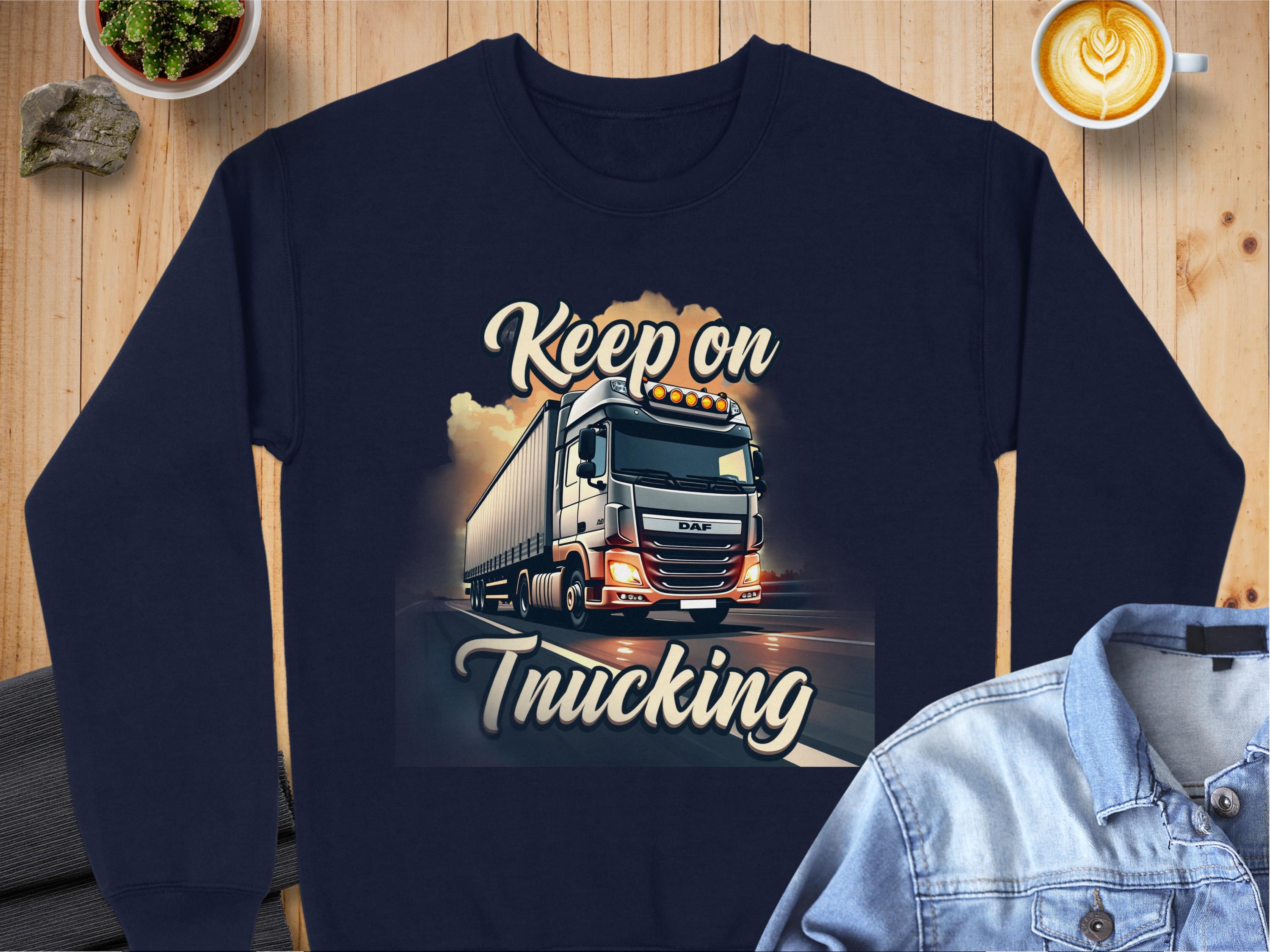 Garment Graphics navy sweatshirt features a large truck with Keep On Trucking text. It pairs well with denim shirts and is set against a wooden surface. With unisex sizing, its perfect for anyone who loves adventure and coffee.