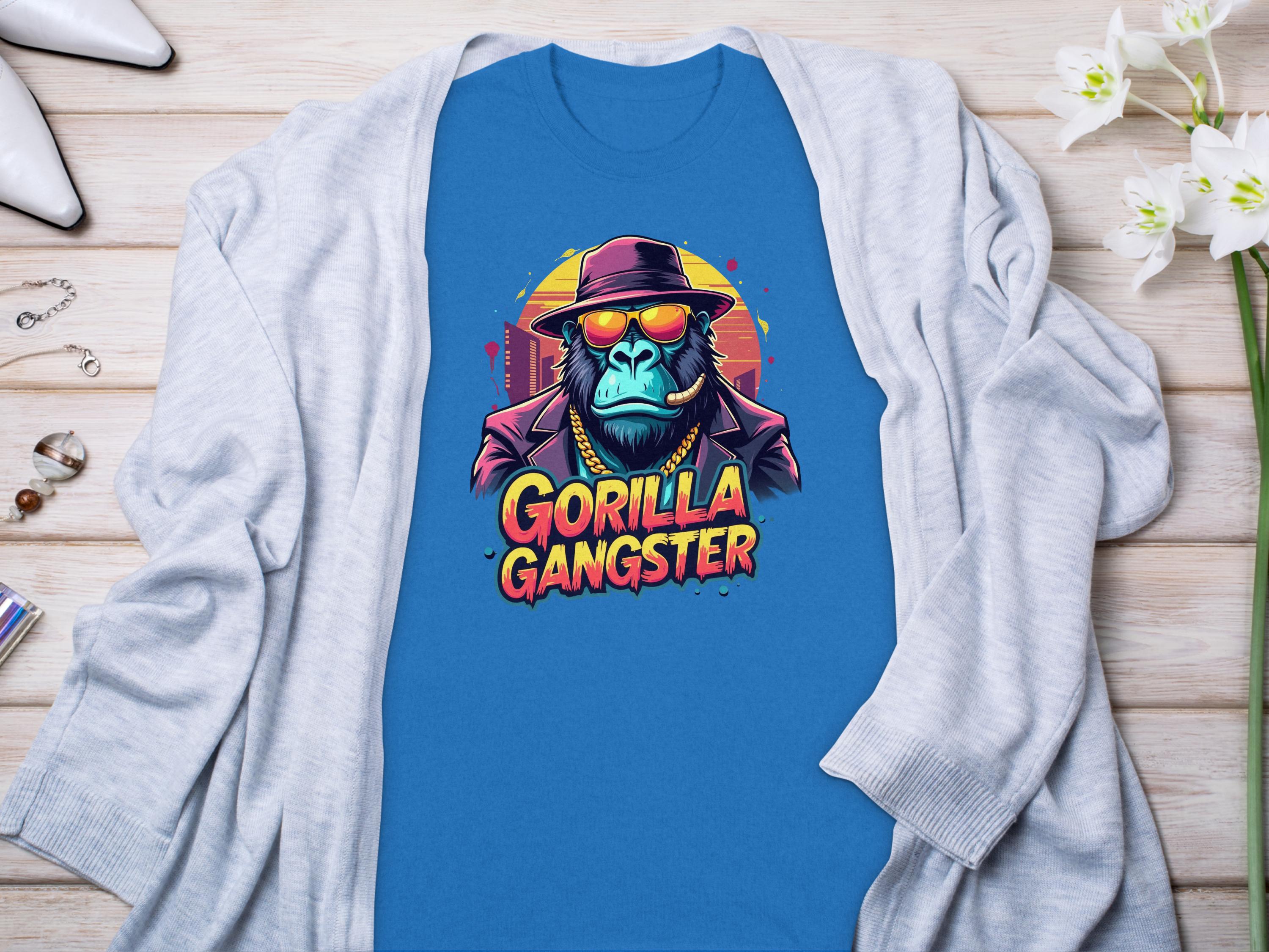 A Garment Graphics blue t-shirt with a Gorilla Gangster design shows a gorilla in a hat and sunglasses. The shirt is displayed on wood, accented by a light gray cardigan and white flowers.