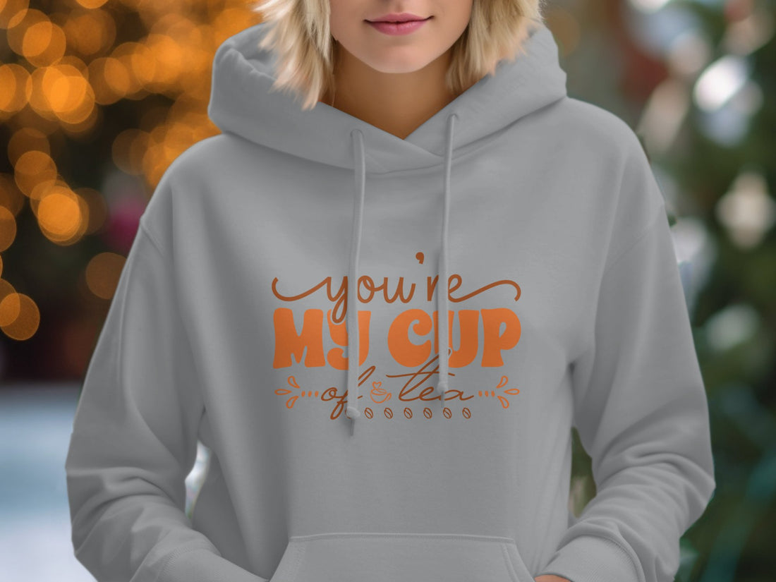 Garment Graphics white hoodie features a youre my cup of tea design in brown, displayed alongside a straw hat, sunglasses, spiral notebook, map, brown sandals, woven bag, and white camera on wood—a perfect setup for capturing cozy moments.