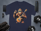 A navy T-shirt from Garment Graphics features a gorilla in sunglasses, jamming on an electric guitar, and flashing a peace sign. Ideal for music lovers, its surrounded by a smartphone, smartwatch, Bluetooth speaker, shoes, and dumbbells on a gray surface.