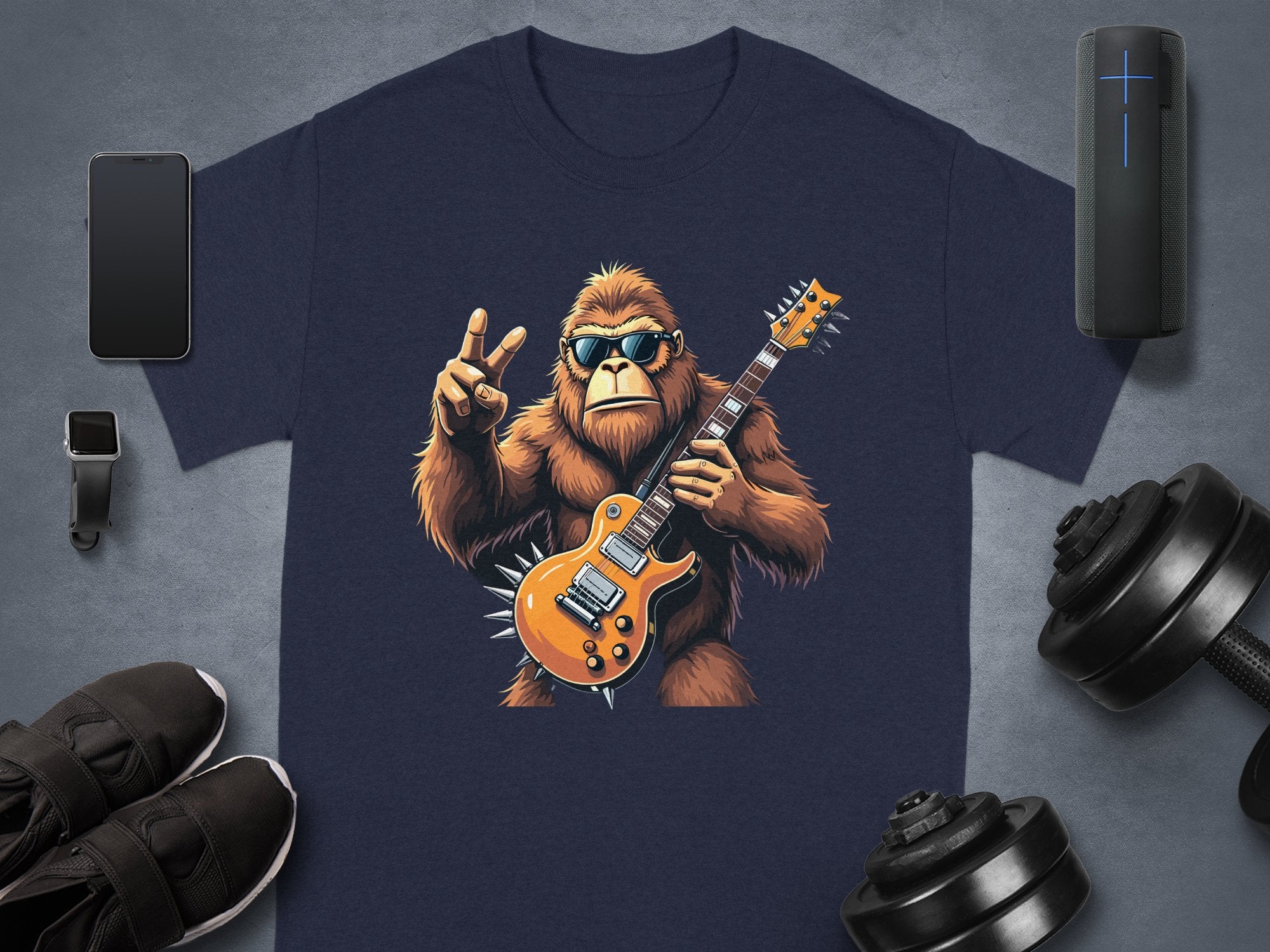 A navy T-shirt from Garment Graphics features a gorilla in sunglasses, jamming on an electric guitar, and flashing a peace sign. Ideal for music lovers, its surrounded by a smartphone, smartwatch, Bluetooth speaker, shoes, and dumbbells on a gray surface.