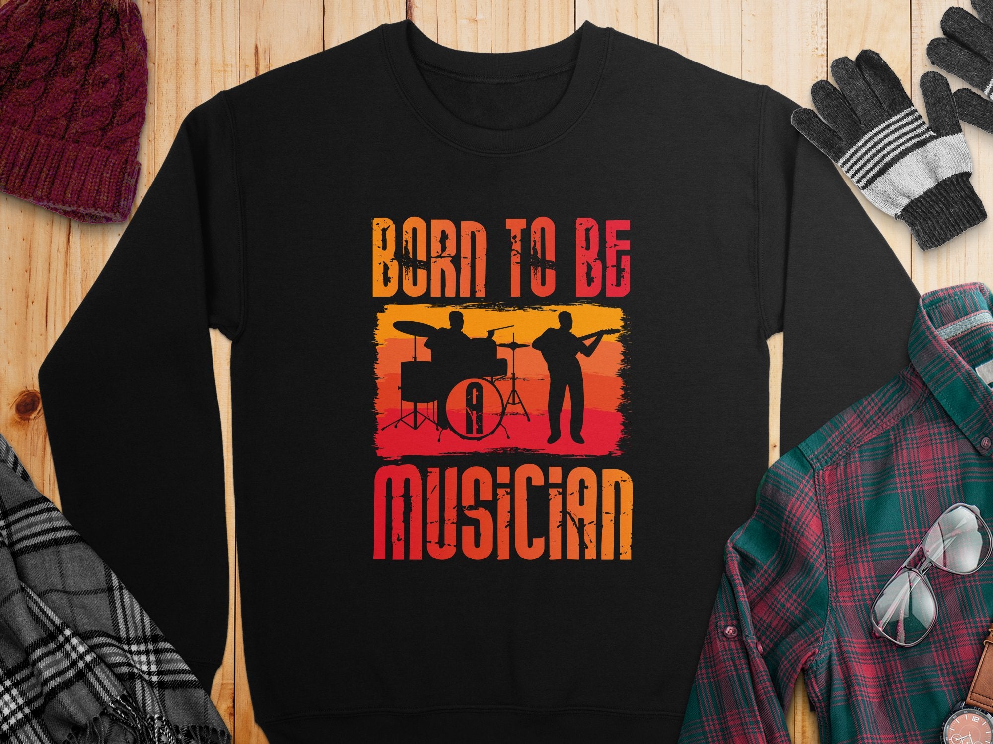 A black unisex sweatshirt from Garment Graphics lies on a wooden surface, ideal for music lovers. It showcases drummer and guitarist silhouettes with Born to Be Musician in gradient orange, red, and yellow. A plaid shirt, gloves, and knit hat surround it.