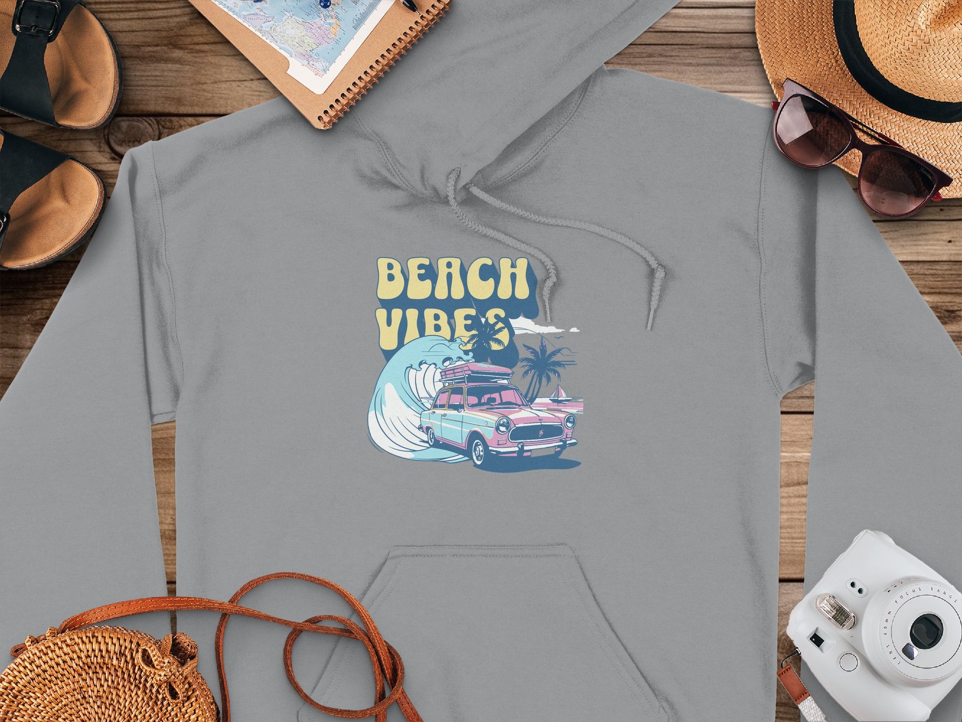 Garment Graphics presents a gray hoodie with a retro beach style, showcasing a Beach Vibes design featuring a pink car, surfboard, and palm trees. Laid on wood, its styled with a straw hat, sunglasses, sandals, notebook, and camera—ideal for vibrant hoodie lovers!.