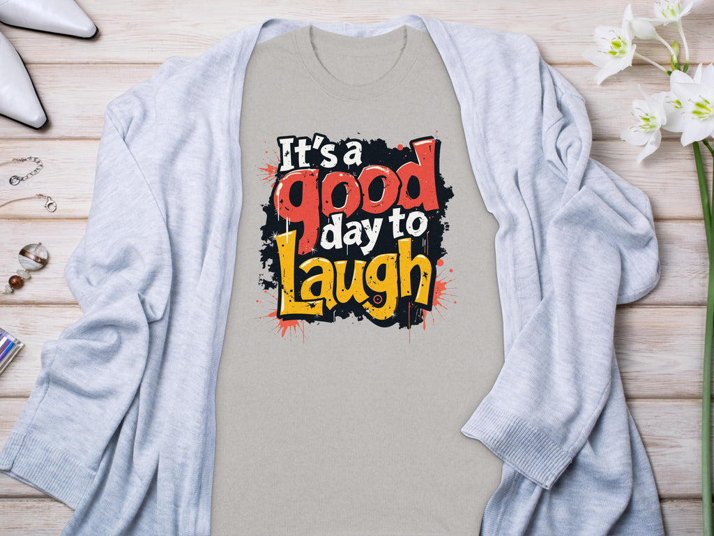 A vibrant gray Its a Good Day to Laugh t-shirt by Garment Graphics is displayed flat on wood, surrounded by a light gray cardigan. White flowers, a perfume bottle, and jewelry beautifully enhance this cheerful setup.