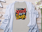 A vibrant gray Its a Good Day to Laugh t-shirt by Garment Graphics is displayed flat on wood, surrounded by a light gray cardigan. White flowers, a perfume bottle, and jewelry beautifully enhance this cheerful setup.