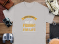 A Garment Graphics light gray fishing T-shirt with Father and Son Fishing Partners For Life in bold orange and white hues lies on a wooden surface, symbolizing family bonding. Nearby are a camera, binoculars, watch, rope, and leather pouch.