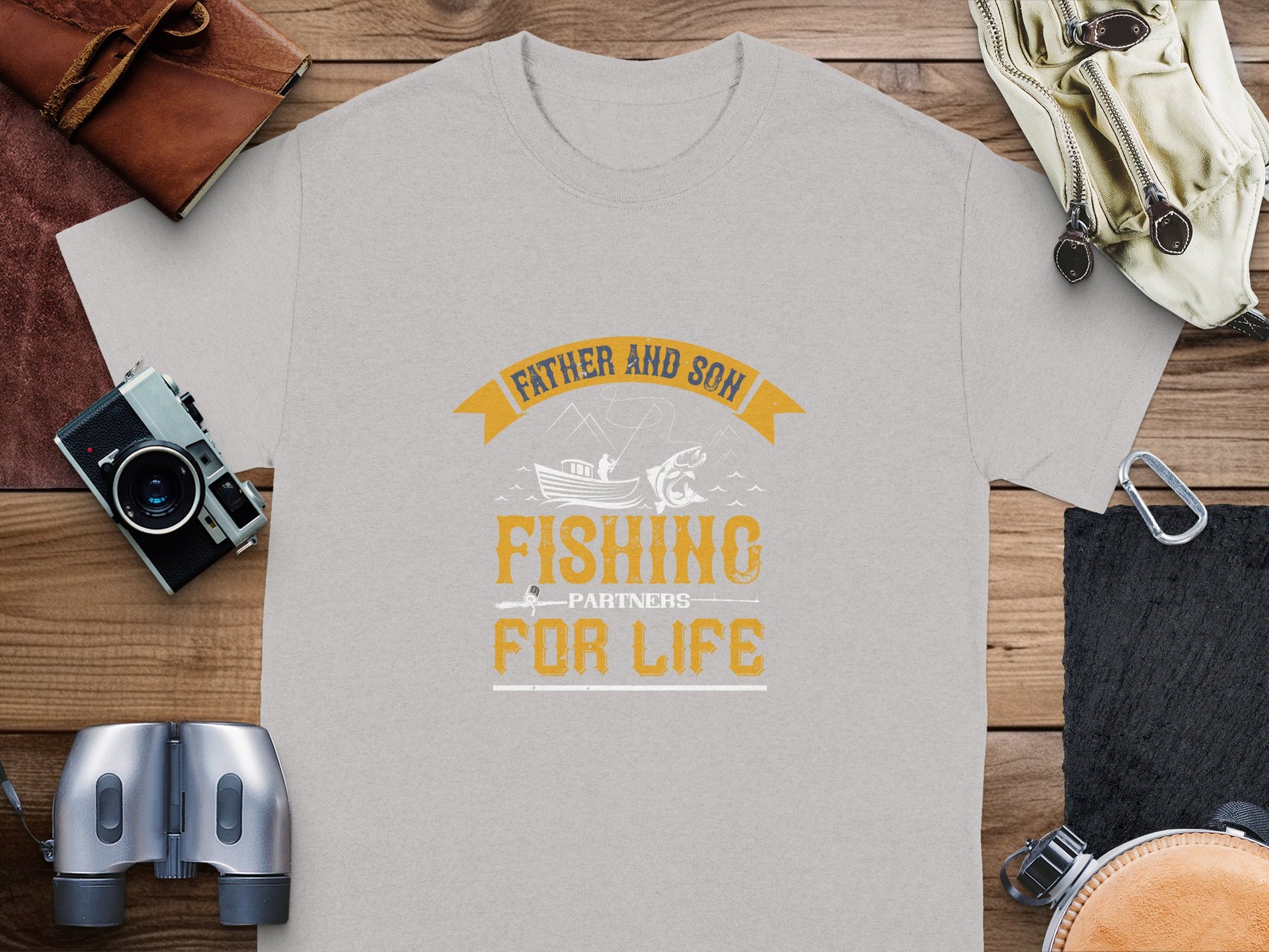 A Garment Graphics light gray fishing T-shirt with Father and Son Fishing Partners For Life in bold orange and white hues lies on a wooden surface, symbolizing family bonding. Nearby are a camera, binoculars, watch, rope, and leather pouch.