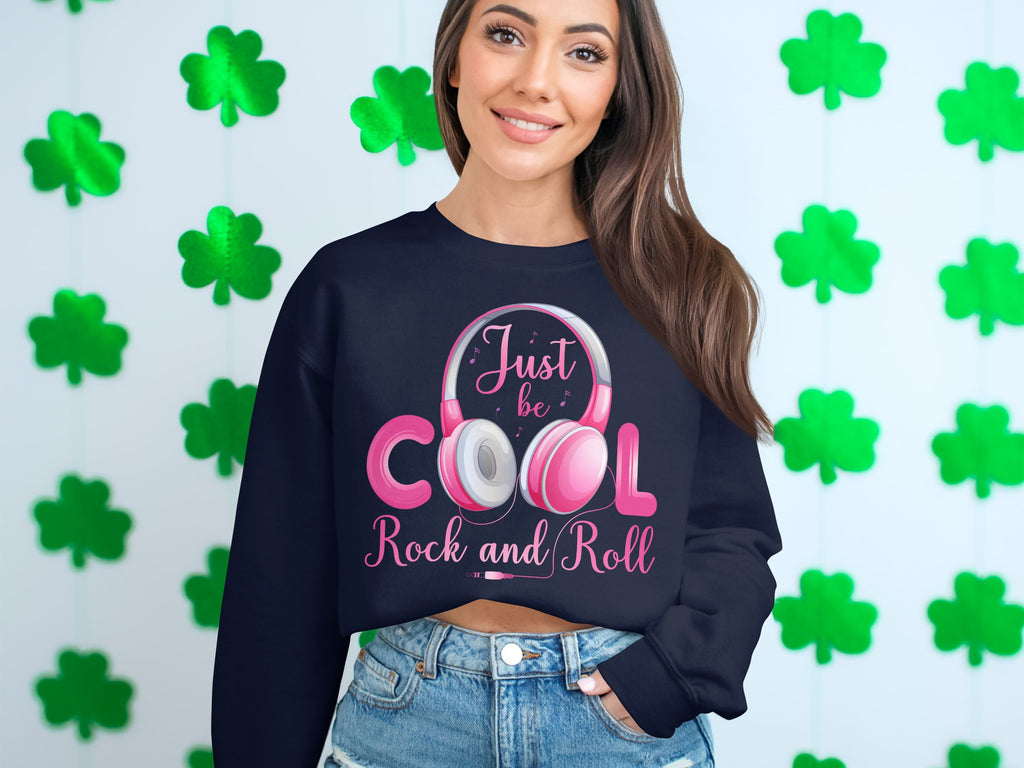 A woman stands before a wall of green shamrocks, wearing a dark Garment Graphics sweatshirt with Just be COOL Rock and Roll and pink headphones, perfect for music lovers. She pairs it with denim shorts, smiling warmly and capturing the essence of fashion enthusiasts everywhere.