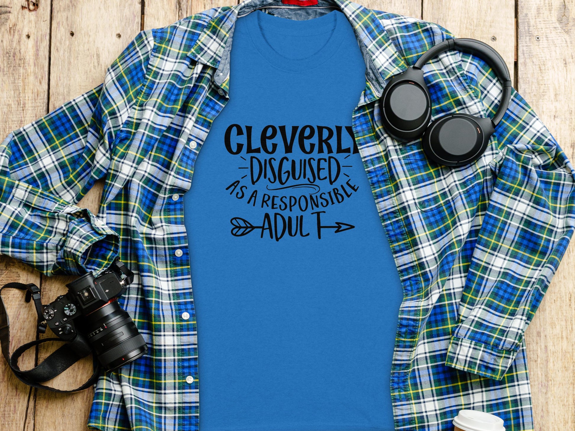 For a casual outing, Garment Graphics classic fit blue tee with Cleverly Disguised as a Responsible Adult pairs perfectly with a green and blue plaid shirt. A black camera and headphones rest beside it on a wooden surface, creating an effortlessly cool ensemble.