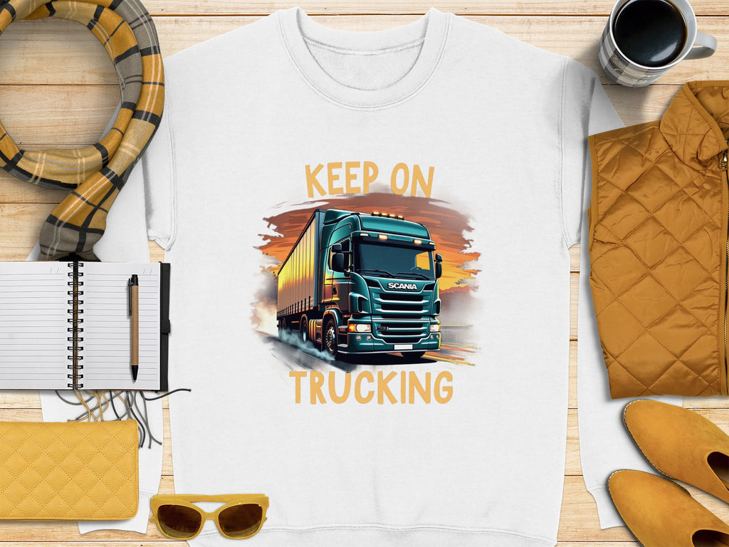 Flat lay of Garment Graphics white KEEP ON TRUCKING tee featuring a large truck graphic. Ideal for truck lovers, its styled with a scarf, mug, yellow purse and sunglasses, notebook with pen, and yellow quilted jacket on a wooden surface.