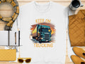 Flat lay of Garment Graphics white KEEP ON TRUCKING tee featuring a large truck graphic. Ideal for truck lovers, its styled with a scarf, mug, yellow purse and sunglasses, notebook with pen, and yellow quilted jacket on a wooden surface.
