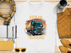 Flat lay of Garment Graphics white KEEP ON TRUCKING tee featuring a large truck graphic. Ideal for truck lovers, its styled with a scarf, mug, yellow purse and sunglasses, notebook with pen, and yellow quilted jacket on a wooden surface.