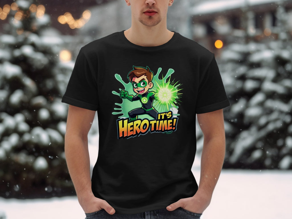 A person stands in the snow wearing a vibrant black Garment Graphics T-shirt featuring a cartoon character with a glowing green orb and the text Its Hero Time! Snow-covered trees and blurred lights provide the perfect backdrop for this superhero graphic print tee.