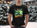 A person stands in the snow wearing a vibrant black Garment Graphics T-shirt featuring a cartoon character with a glowing green orb and the text Its Hero Time! Snow-covered trees and blurred lights provide the perfect backdrop for this superhero graphic print tee.