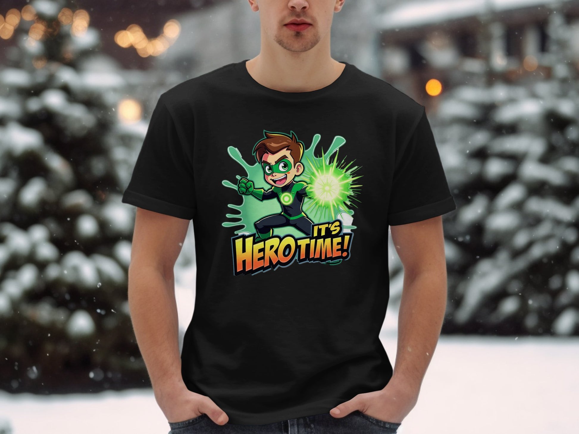 A person stands in the snow wearing a vibrant black Garment Graphics T-shirt featuring a cartoon character with a glowing green orb and the text Its Hero Time! Snow-covered trees and blurred lights provide the perfect backdrop for this superhero graphic print tee.