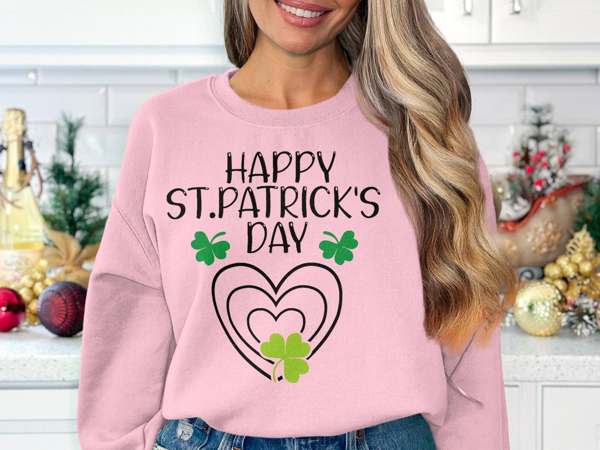 A person in a Garment Graphics sweatshirt featuring Happy St. Patricks Day and a clover design stands in a kitchen adorned with gold and red ornaments, enhancing the holiday cheer.