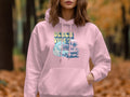 A person wears a vibrant Garment Graphics hoodie with a surfboard design, featuring a car, palm trees, ocean waves, and the text BEACH VIBES, set against an autumn-colored forest backdrop.